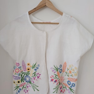 May include: A white short-sleeved top featuring a round neckline and a front opening. The garment is decorated with two symmetrical, colourful floral embroidery patterns on the lower front panels, showcasing various flowers in shades of pink, blue, yellow, and green.