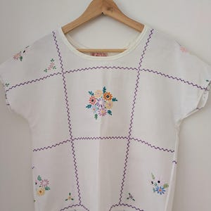 May include: A white short-sleeved top featuring a grid pattern created with purple zigzag embroidery stitches. The garment is decorated with small, colourful floral embroidery motifs in shades of pink, yellow, blue, and green. A label reads Made by Miss Jonas.