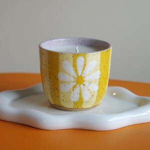 May include: A yellow ceramic candle vessel with a white daisy pattern and speckled texture, containing white wax with a single wick, resting on a white, cloud-shaped ceramic tray. The vessel features vertical yellow stripes and a light purple rim.