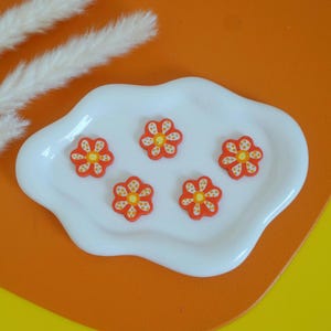 May include: Five orange flower-shaped embellishments with yellow centers and white accents are arranged on a white, cloud-shaped dish. The dish sits on an orange surface, with a yellow background. Decorative floral elements.