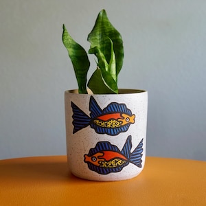 May include: A cylindrical white ceramic planter with a speckled finish, featuring two hand-painted fish illustrations. Each fish has a bright orange body, a yellow spotted belly, and dark blue fins. A green snake plant grows from the planter.
