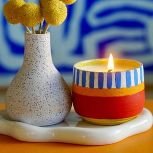 May include: A lit candle in a colorful ceramic container with blue, orange, and red stripes. A white speckled vase with yellow flowers sits next to the candle on a white, wavy tray. The background is a blue and white abstract design.