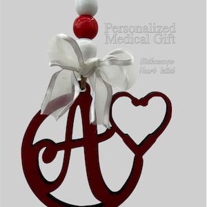 May include: A burgundy wooden ornament with the letter "A" and a heart-shaped stethoscope design. The ornament is adorned with a white ribbon and a string of white and red beads. Text on the image reads "Personalized Medical Gift" and "Stethoscope Heart Initial".