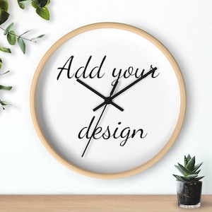 Op de afbeelding: Een ronde wandklok met een licht houten frame en een witte wijzerplaat. De klokwijzers zijn zwart en de tekst "Add your design" staat in zwart schrift op de wijzerplaat. Een kleine vetplant is zichtbaar.