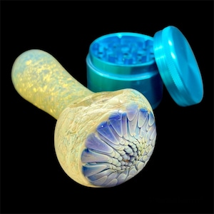 May include: A glass pipe with a blue and white swirl design. The pipe is made of clear glass with a yellow tint. The pipe is sitting next to a blue metal grinder.