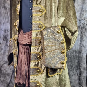 May include: An olive green velvet coat featuring gold-toned buttons and grey paisley patterned panels on the front and cuffs. The coat is worn over a dark blue shirt and a brown fabric sash, with gold trim bordering the patterned sections.