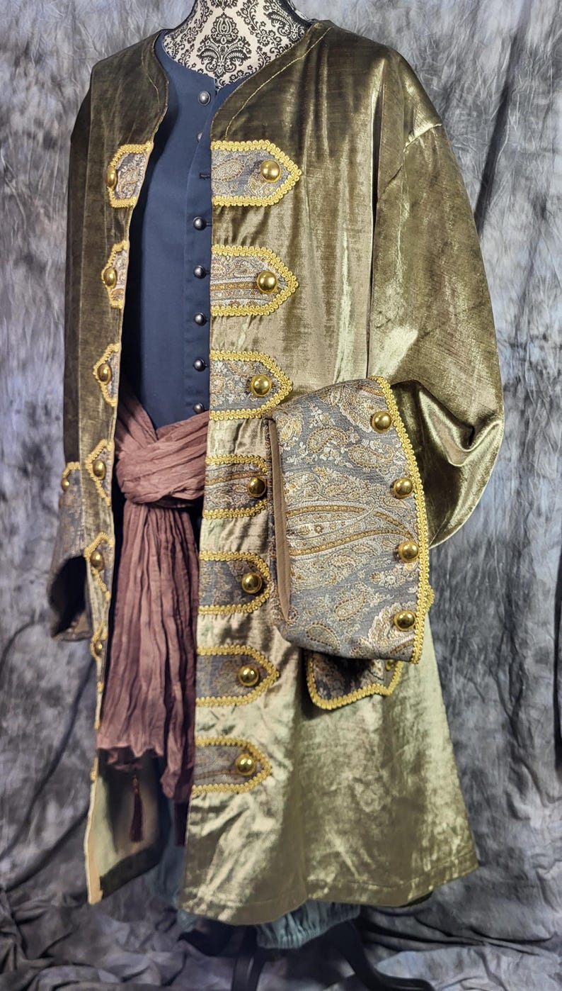 May include: An olive green velvet coat featuring gold-toned buttons and grey paisley patterned panels on the front and cuffs. The coat is worn over a dark blue shirt and a brown fabric sash, with gold trim bordering the patterned sections.