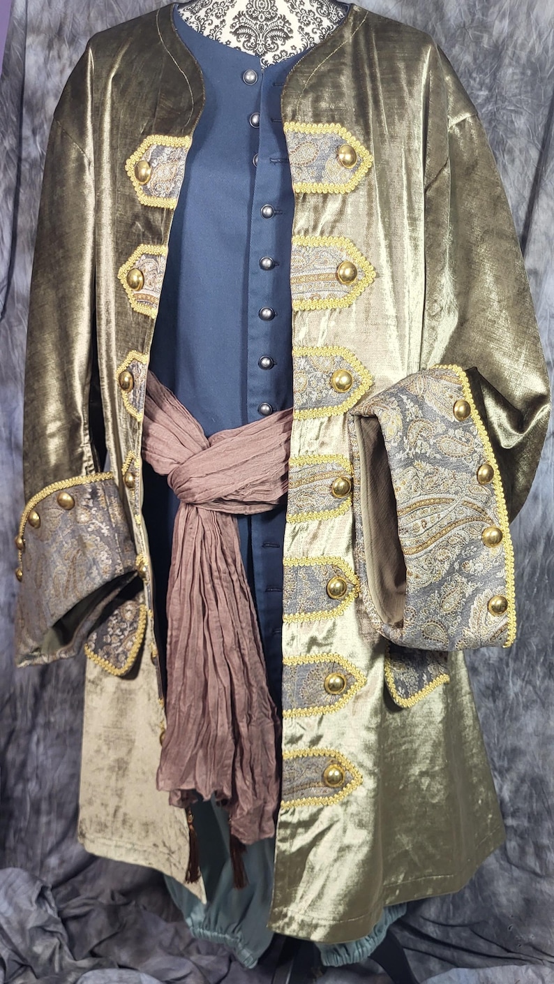 May include: A metallic olive green long coat with a dark blue inner tunic and a brown sash. The coat features gold-trimmed grey paisley panels, brass-colored buttons, and wide cuffs. The inner tunic has a row of small silver-colored buttons.