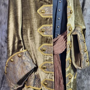 May include: An olive green velvet coat featuring gold-toned buttons and decorative panels with a grey and gold paisley pattern. The coat is worn over a dark blue shirt and a brown fabric sash, with wide cuffs that match the paisley panels.