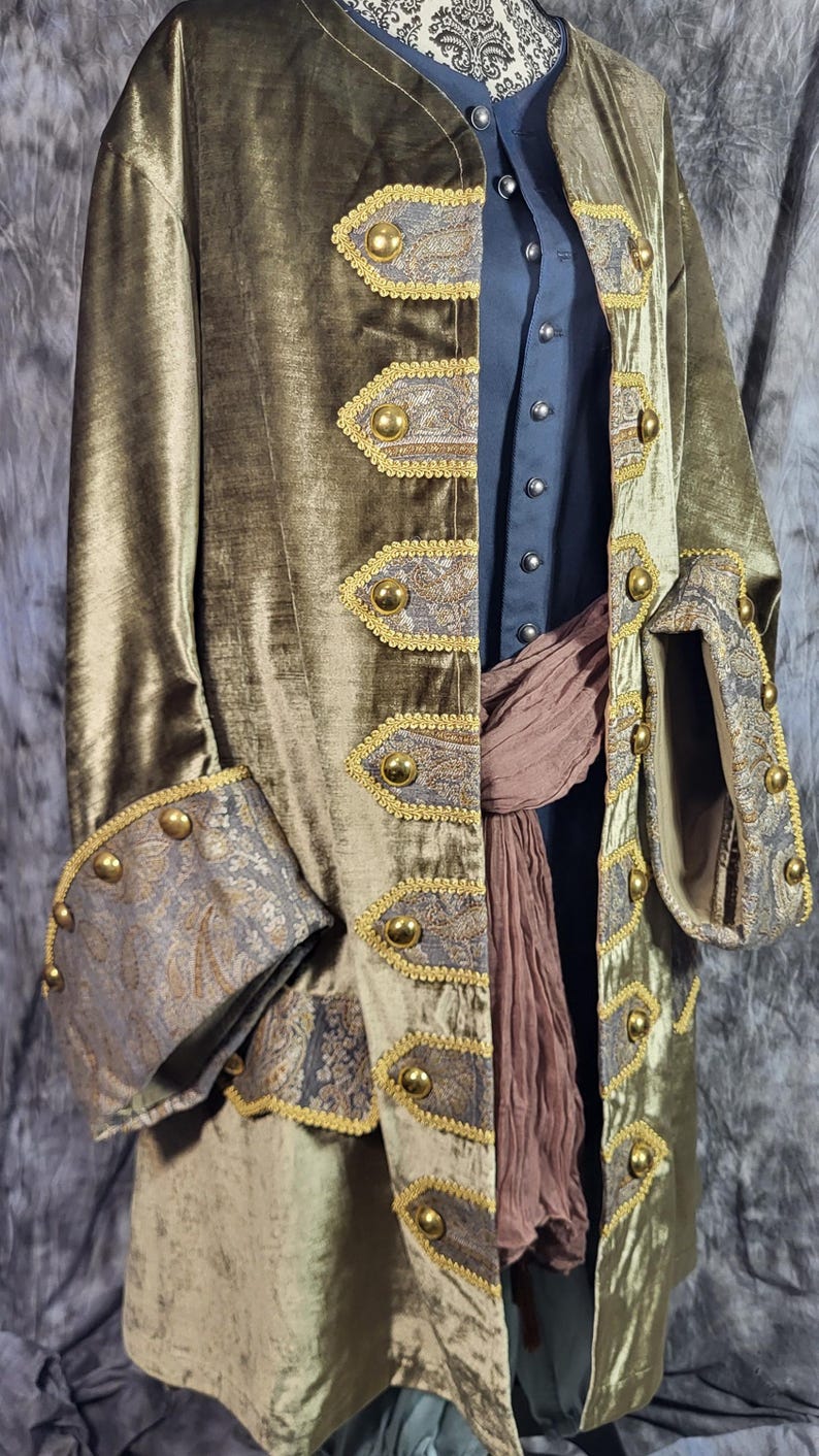 May include: An olive green velvet coat featuring gold-toned buttons and decorative panels with a grey and gold paisley pattern. The coat is worn over a dark blue shirt and a brown fabric sash, with wide cuffs that match the paisley panels.