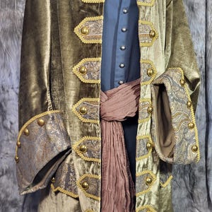 May include: An olive green velvet coat featuring gold-toned buttons and decorative paisley-patterned panels along the front opening and cuffs. The coat is worn over a dark blue button-up shirt and a textured brown sash tied around the waist.