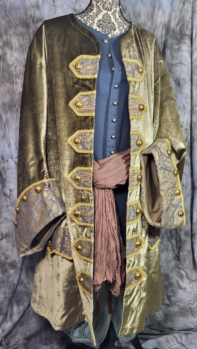 May include: An olive green velvet coat featuring gold-toned buttons and decorative paisley-patterned panels along the front opening and cuffs. The coat is worn over a dark blue button-up shirt and a textured brown sash tied around the waist.