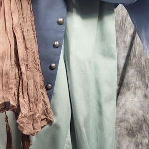 May include: A light blue long-sleeved shirt worn under a dark blue vest with silver-toned buttons. A light brown crinkled scarf with tassels hangs over the shoulder. The outfit includes light green trousers with elasticized cuffs at the ankles.