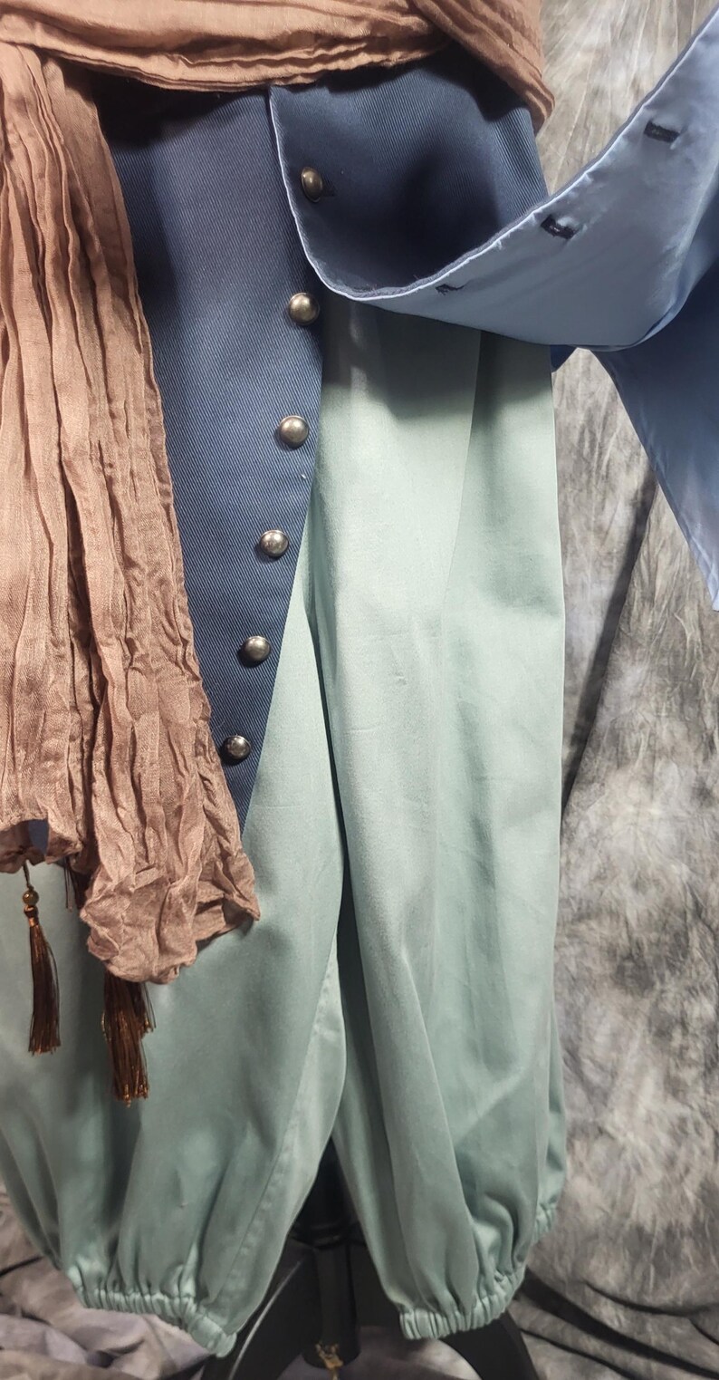 May include: A light blue long-sleeved shirt worn under a dark blue vest with silver-toned buttons. A light brown crinkled scarf with tassels hangs over the shoulder. The outfit includes light green trousers with elasticized cuffs at the ankles.