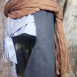 May include: A light blue sleeveless top with a side tie closure, layered under a dark charcoal gray vest with a structured hem. A long, crinkled brown scarf with tassel ends is wrapped around the waist, adding texture to the layered garment ensemble.