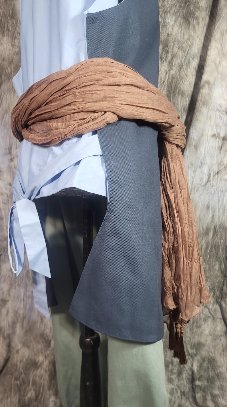 May include: A light blue sleeveless top with a side tie closure, layered under a dark charcoal gray vest with a structured hem. A long, crinkled brown scarf with tassel ends is wrapped around the waist, adding texture to the layered garment ensemble.