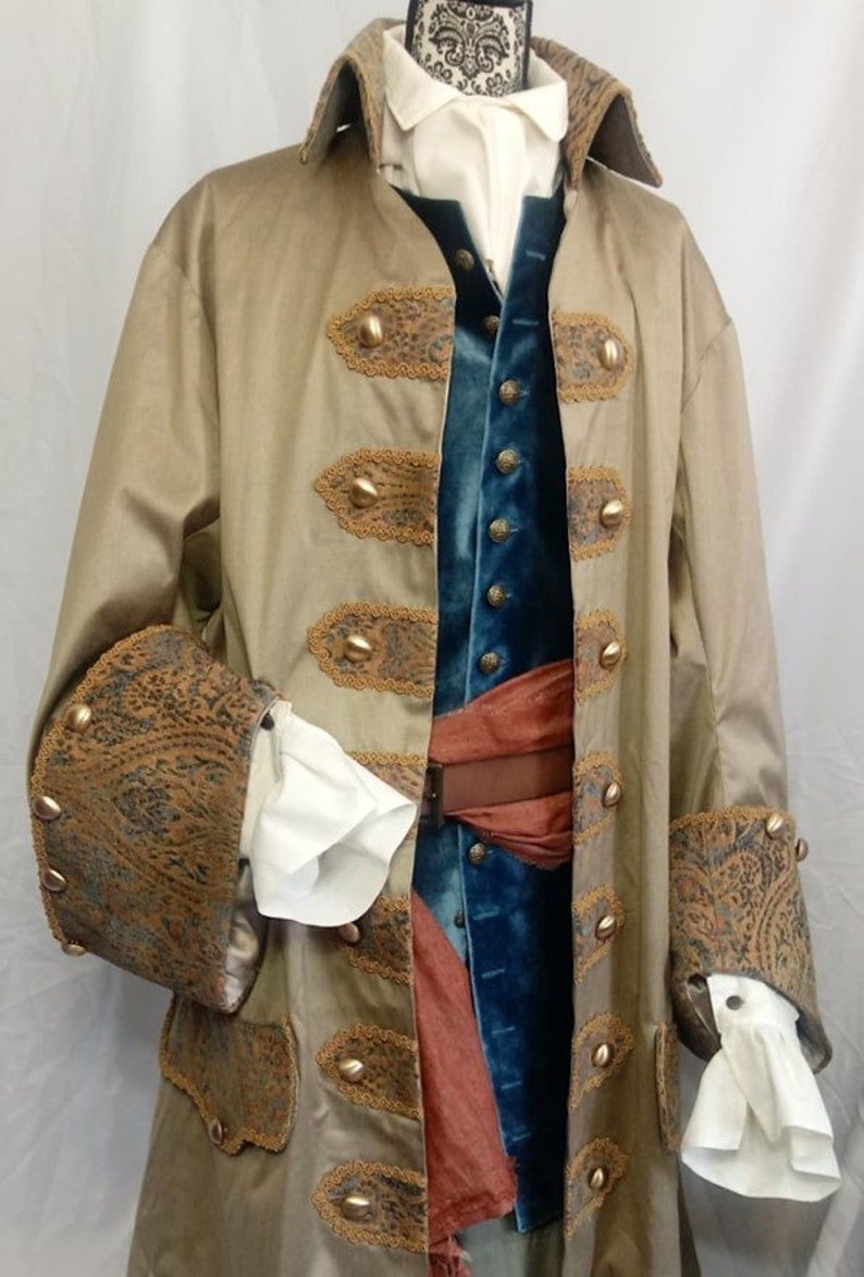 May include: A tan long coat with wide cuffs and pocket flaps featuring a dark paisley pattern, worn over a blue velvet vest and a white shirt with ruffled cuffs. A brown belt and a rust-colored sash are layered over the blue vest.