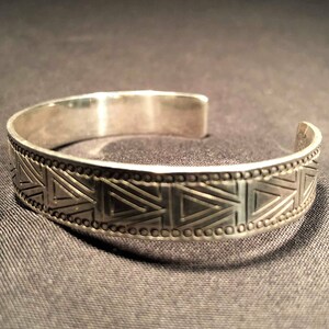 May include: Silver cuff bracelet featuring a geometric triangle design. The open-ended bracelet is made of a shiny metal. The triangles are arranged in a repeating pattern along the band, with a dotted border.