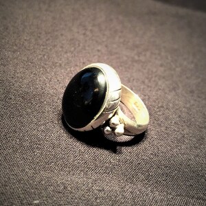 Peut inclure: Une bague en argent avec une pierre noire ovale. La bague a un anneau texturé et de petits ornements ronds près de la pierre. La bague est posée sur un fond gris foncé.