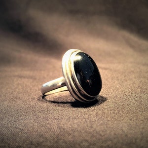 Peut inclure: Une bague en argent ornée d'une grosse pierre noire ovale. La pierre est sertie dans une lunette en argent. La bague a une surface polie et réfléchissante. L'anneau est simple et lisse, de conception classique.