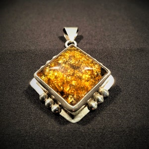 Op de afbeelding: Een zilveren hanger met een vierkante amber steen. De steen is diep goudkleurig en heeft veel insluitsels. De hanger is gezet in een zilveren frame met een diamantvorm.