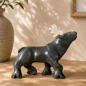 Peut inclure: Une sculpture en pierre polie vert foncé représentant un élan debout avec une finition lisse. L'animal présente un bois de couleur crème clair distinct fixé sur sa tête, contrastant avec le corps sombre et les quatre pattes robustes.