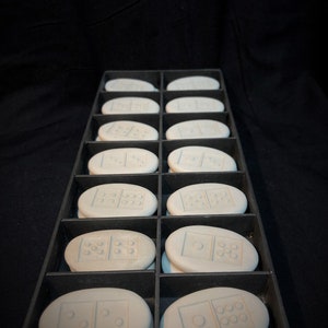 Puede incluir: Un juego de 12 fichas de dominó blancas con puntos negros, dispuestas en una bandeja negra. Las fichas de dominó son de forma ovalada y tienen un acabado liso y mate.