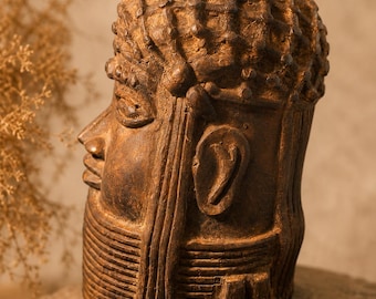 May include: A detailed, brown sculpture of a head, showcasing intricate carvings and textures. The sculpture features a prominent nose, defined facial features, and a textured hairstyle. The piece is set on a stone surface, suggesting an ancient or historical theme.
