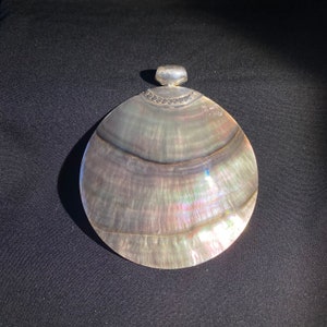 Puede incluir: Un colgante de plata con una gran concha de abulón redonda e iridiscente. La concha tiene un borde plateado.