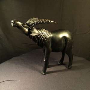 Puede incluir: Una escultura de madera negra de un antílope con la cabeza echada hacia atrás y los cuernos curvados hacia adelante. La escultura está tallada con intrincados detalles y tiene un acabado liso y pulido.