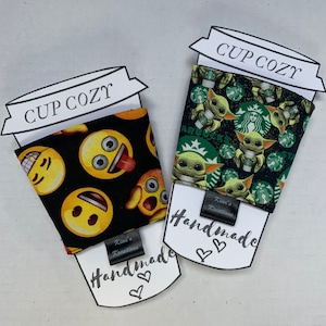May include: Two fabric cup cozies. One features yellow emoji faces on a black background. The other has a pattern of Baby Yoda and Starbucks logos on a black background. Each cozy has a tag that says "Handmade". The top of each cozy has a paper cutout that says "Cup Cozy".