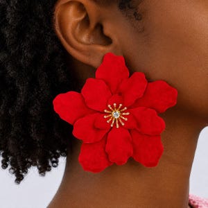 May include: A large, vibrant red flower-shaped earring featuring layered, matte-finish petals. The center of the flower is adorned with a gold-toned starburst design, accented by a single clear, sparkling crystal at the very middle of the piece.