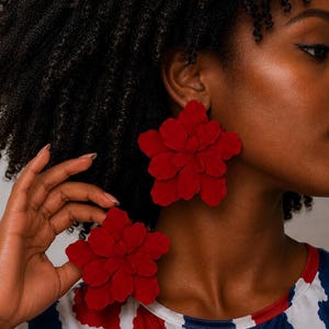 May include: A pair of large, deep red flower-shaped earrings with layered, petal-like components. The earrings feature a matte, textured finish and a bold, oversized design that covers a significant portion of the earlobe area.