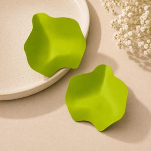 May include: A pair of lime green earrings with an irregular, wavy, organic shape. The earrings feature a smooth, matte finish and a curved, petal-like design that creates subtle shadows across the surface of each piece.