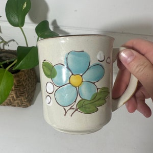 Puede incluir: Una taza de cer&aacute;mica blanca con una flor azul y hojas verdes pintadas en el lateral. La flor tiene un centro amarillo.