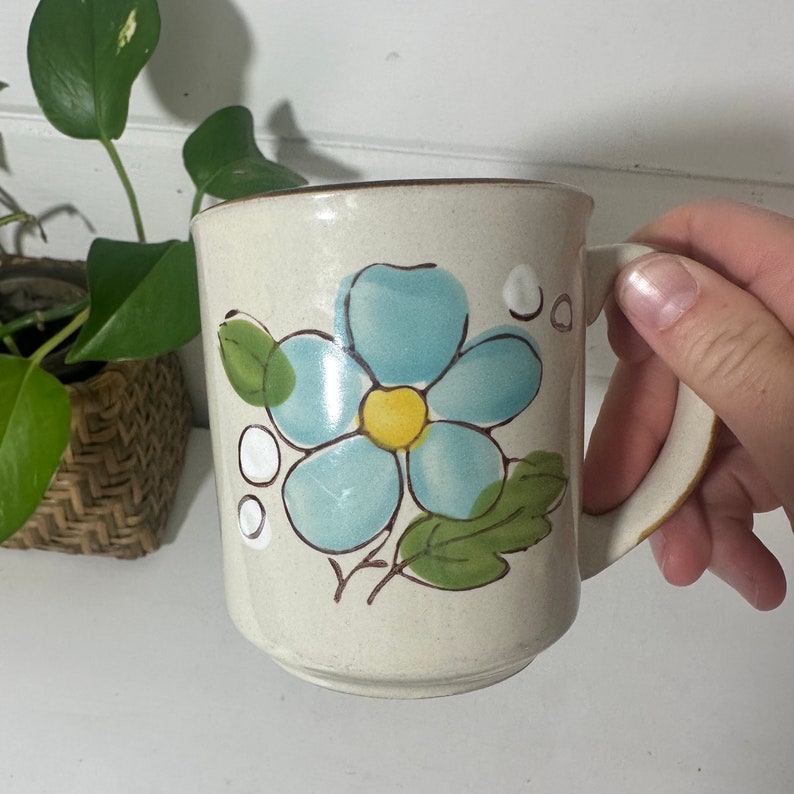 Puede incluir: Una taza de cer&aacute;mica blanca con una flor azul y hojas verdes pintadas en el lateral. La flor tiene un centro amarillo.