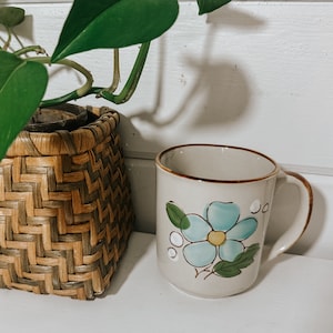 Puede incluir: Una taza de cer&aacute;mica blanca con un dise&ntilde;o de flor azul. La taza tiene un borde y un asa marrones. La taza est&aacute; sobre una superficie blanca.