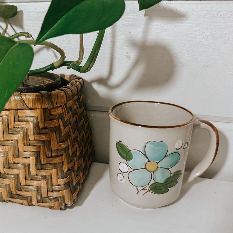 Puede incluir: Una taza de cer&aacute;mica blanca con un dise&ntilde;o de flor azul. La taza tiene un borde y un asa marrones. La taza est&aacute; sobre una superficie blanca.