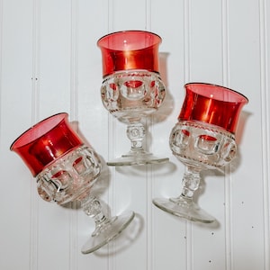 May include: Three vintage wine glasses with ruby red rims and clear, patterned bowls and stems. The glasses are arranged against a white background. The glasses have a classic, elegant design.