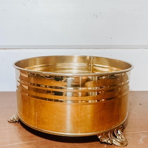 May include: A round, gold-colored metal planter with decorative feet. The planter has a wide, open top and horizontal bands around the sides. The planter is made of a shiny, reflective metal.