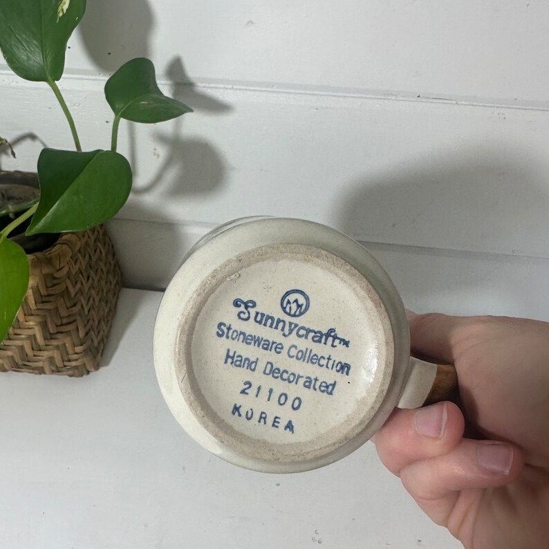 Puede incluir: Una taza de cer&aacute;mica blanca con un dise&ntilde;o azul y blanco. La taza tiene el texto "Sunnycraft Stoneware Collection Hand Decorated 21100 KOREA" en la parte inferior.