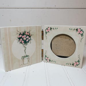 Può includere: Una cornice doppia incernierata con sfondo a righe color crema e beige. Il pannello sinistro mostra un'illustrazione di un topiario di rose in vaso. Il pannello destro ha un'apertura circolare per una foto, decorata con motivi di rose rosa e viticci.