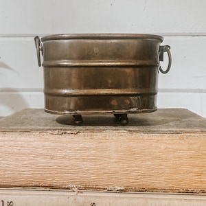 May include: A small, antique copper-colored metal planter with a cylindrical shape. It features two small handles and rests on three small ball feet. The planter is placed on top of a stack of old books.