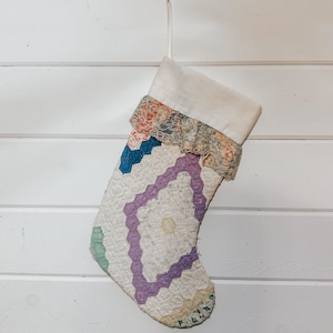 May include: A quilted Christmas stocking with a white cuff and a decorative lace trim. The body of the stocking features a geometric pattern in shades of purple, blue, and green on a white background. The stocking is hanging against a white wooden wall.
