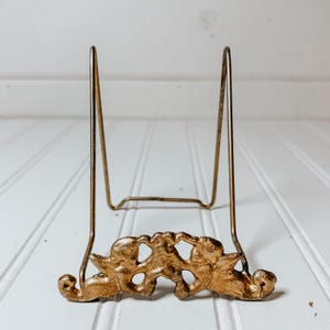 May include: A gold-colored metal display stand with a decorative cherub design at the base. The stand has a simple, angled frame to hold a plate or picture. The cherubs are detailed and add a vintage touch.