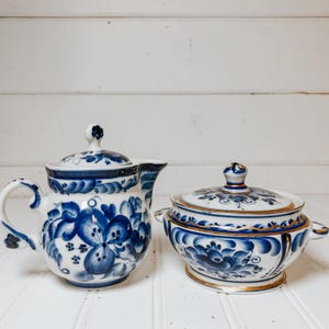 May include: A white ceramic teapot and covered bowl set. The pieces are decorated with blue floral patterns and gold trim. The teapot has a curved handle and spout. The bowl has two small handles. The set is on a white surface.