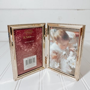 May include: A gold-colored metal double photo frame, open to display a baby photo and a decorative burgundy backing. The frame is 3 1/2 x 5 inches (9 x 13 cm) and has a beaded edge design. The frame is labeled "ROYAL" and "METAL FRAME".