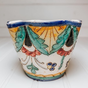 May include: A white ceramic planter with a colorful floral design. The planter features a blue rim, with orange, green, and red floral patterns. The planter is a decorative item for indoor or outdoor use.