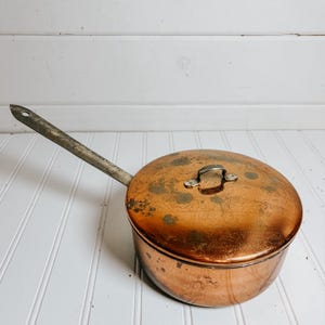 May include: A copper saucepan with a lid and a long, silver-colored handle. The pot has a round shape and a tarnished, aged appearance. The lid features a small handle for easy lifting. The saucepan is likely vintage or antique.
