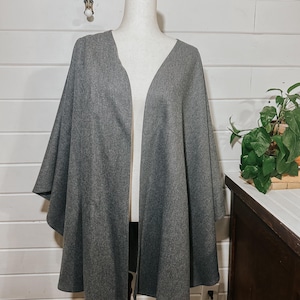 May include: A gray cape-style wrap with a V-neck design, displayed on a mannequin. The wrap has a soft, textured appearance and drapes loosely. The background features a white-painted wood wall and a small plant.